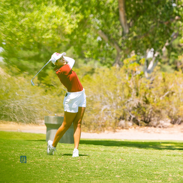 A female golfer in a red shirt and white shorts follows through on a swing in a lush green landscape.