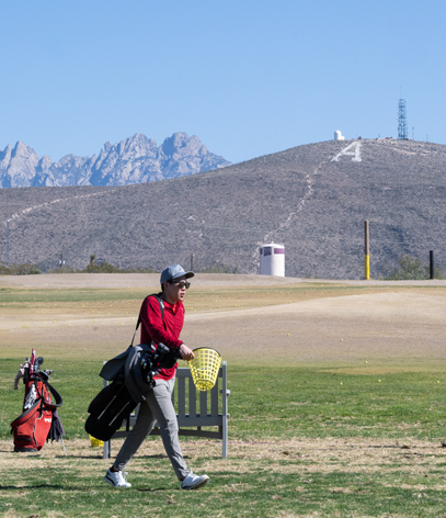 A golfer walks across a driving range with mountains in the background and a large letter "A" on the hillside.